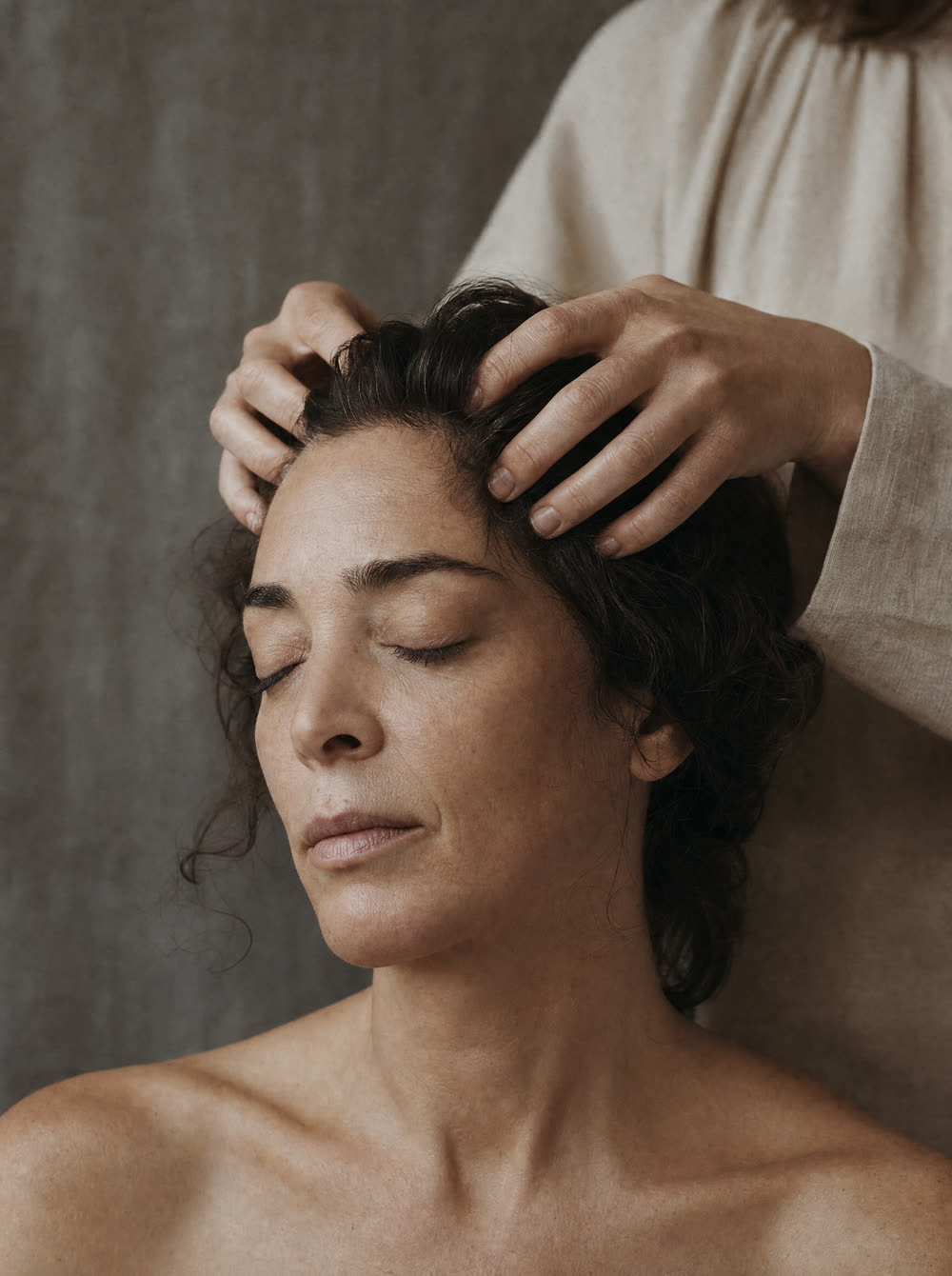A guest with her eyes closed during a scalp massage, a therapist's hands resting at her hairline.