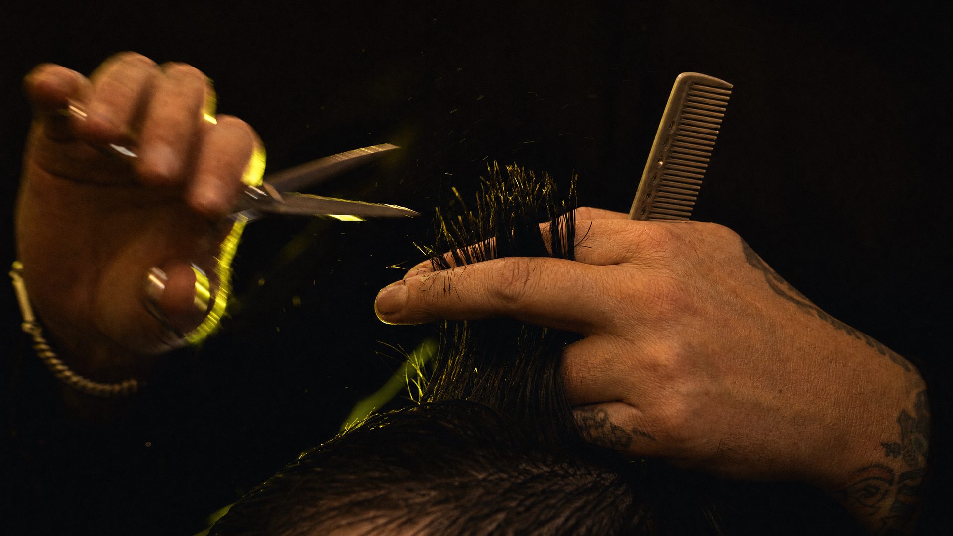 A stylist's hands mid cut, shears open, hair lifted between two fingers, lit by a chartreuse rim light