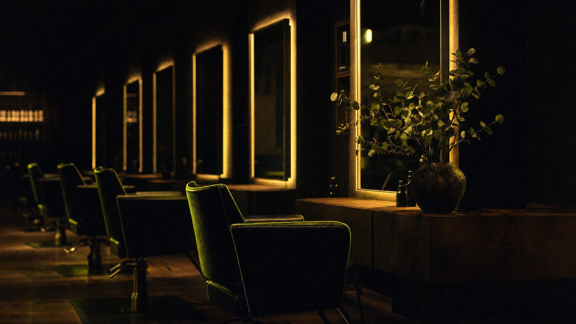 A dark salon at night: a row of green styling chairs and backlit mirrors, eucalyptus in a vase, a single station glowing warm
